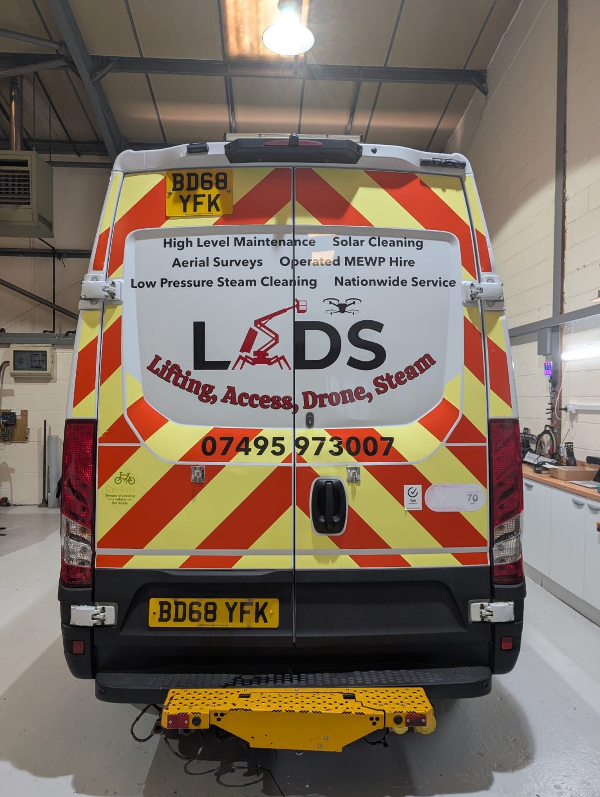 The rear of the LADS van in the workshop, its chevron markings and vinyl graphics listing high level maintenance, solar cleaning, aerial surveys, operated MEWP hire and low pressure steam cleaning